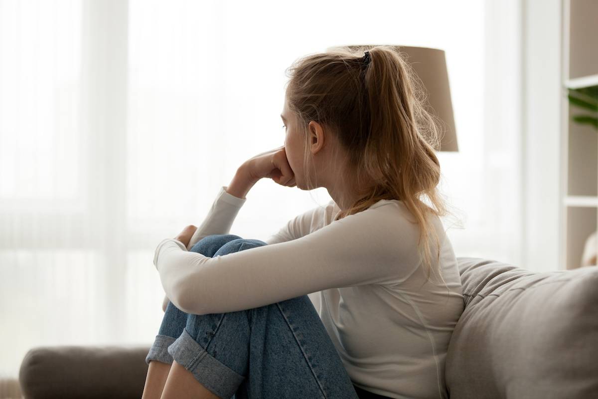 risk factors for addiction Side view young woman looking away at window sitting on couch at home. Frustrated confused female feels unhappy problem in personal life quarrel break